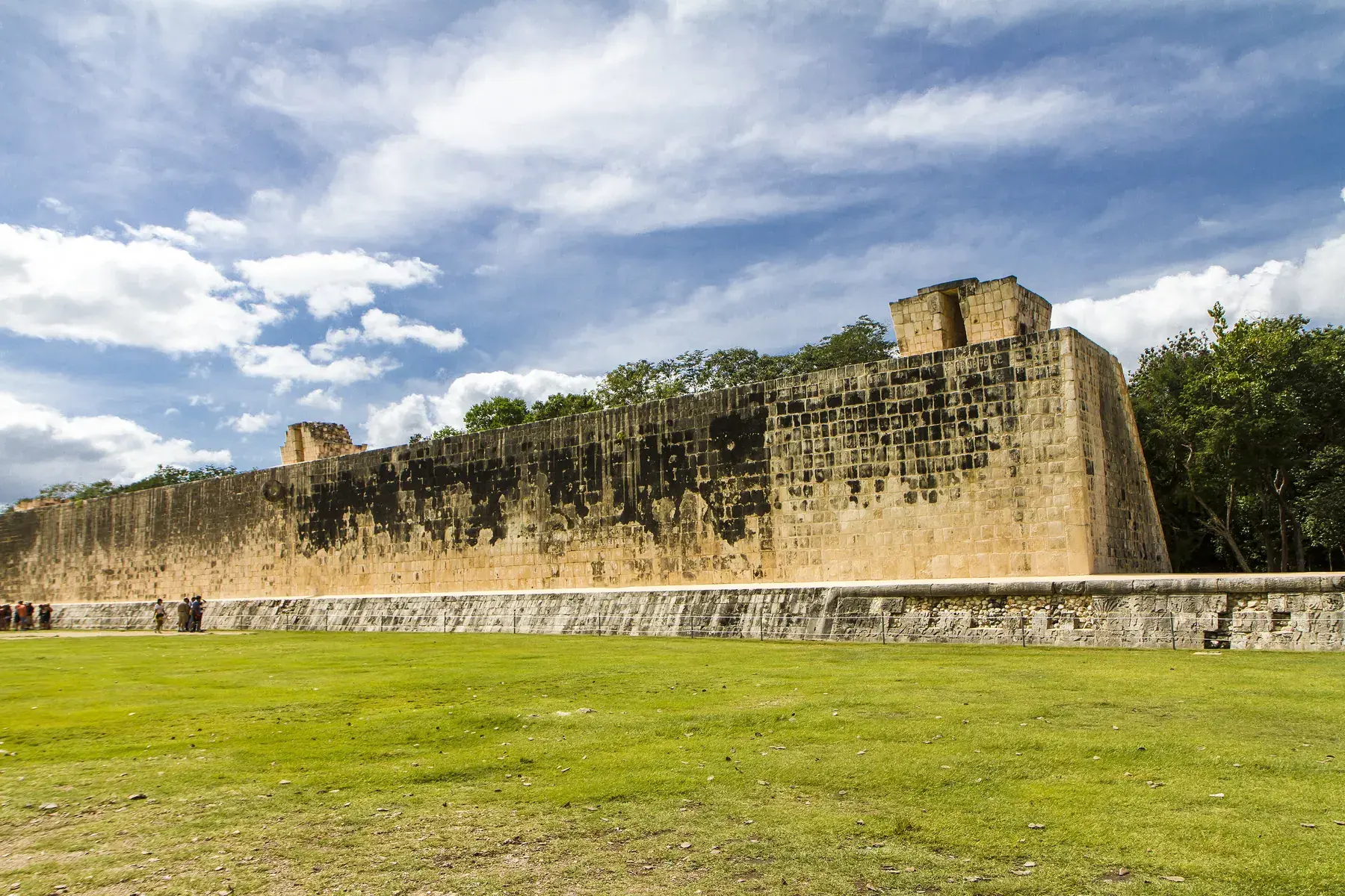 Stone wall of the Great Ball Court at Chichén Itzá, with a lawn and a blue sky with clouds