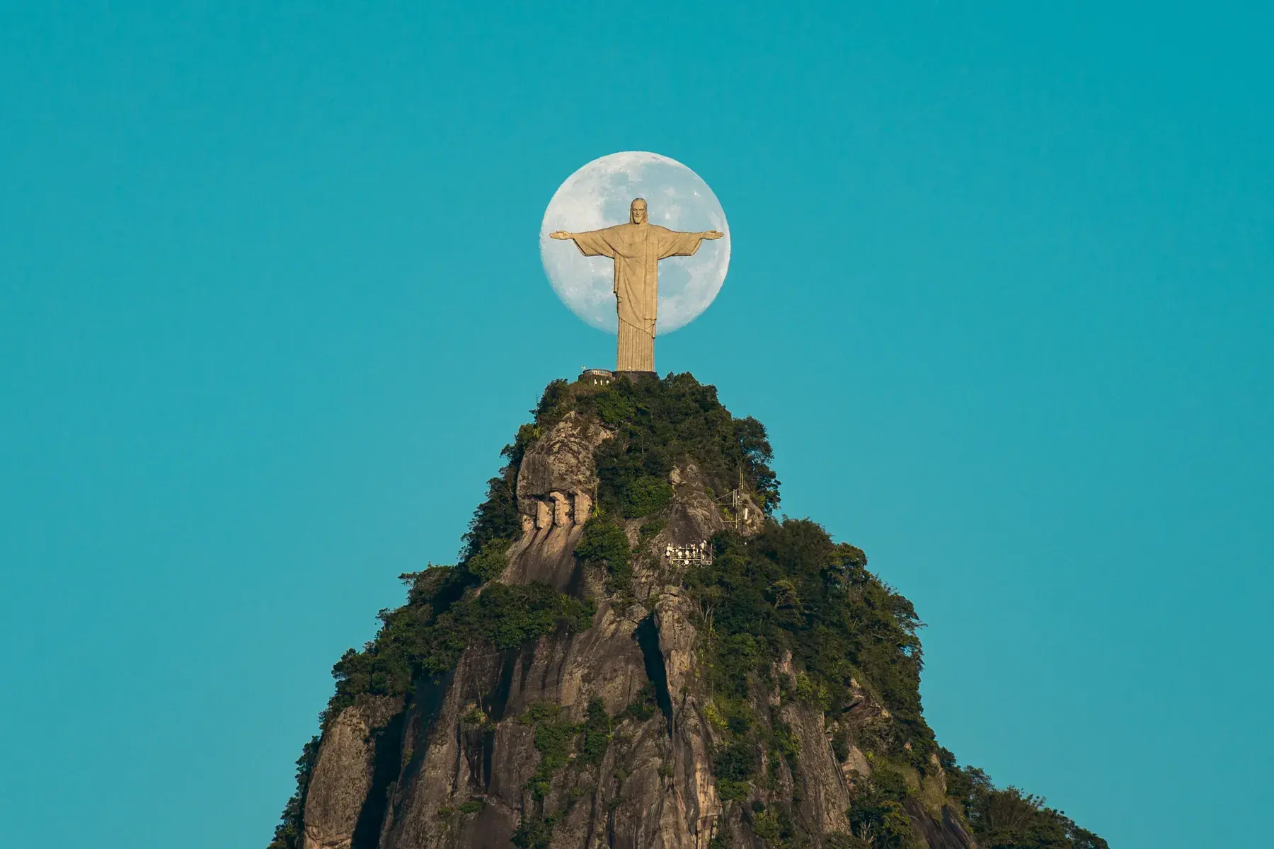 Full moon behind the statue of Christ the Redeemer, atop Corcovado, in a turquoise-blue sky