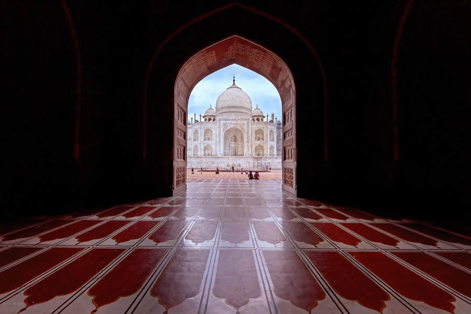 Taj Mahal seen in the distance through a dark arch, with a red sandstone floor in the foreground