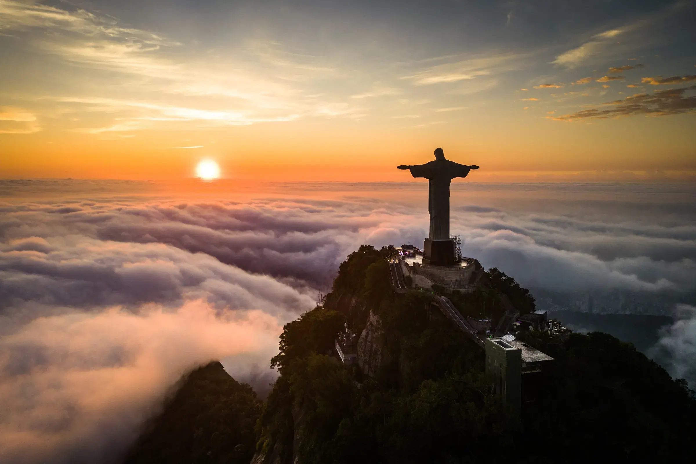 Silhouette of Christ the Redeemer above a sea of golden clouds, with the sun low on the Rio de Janeiro horizon